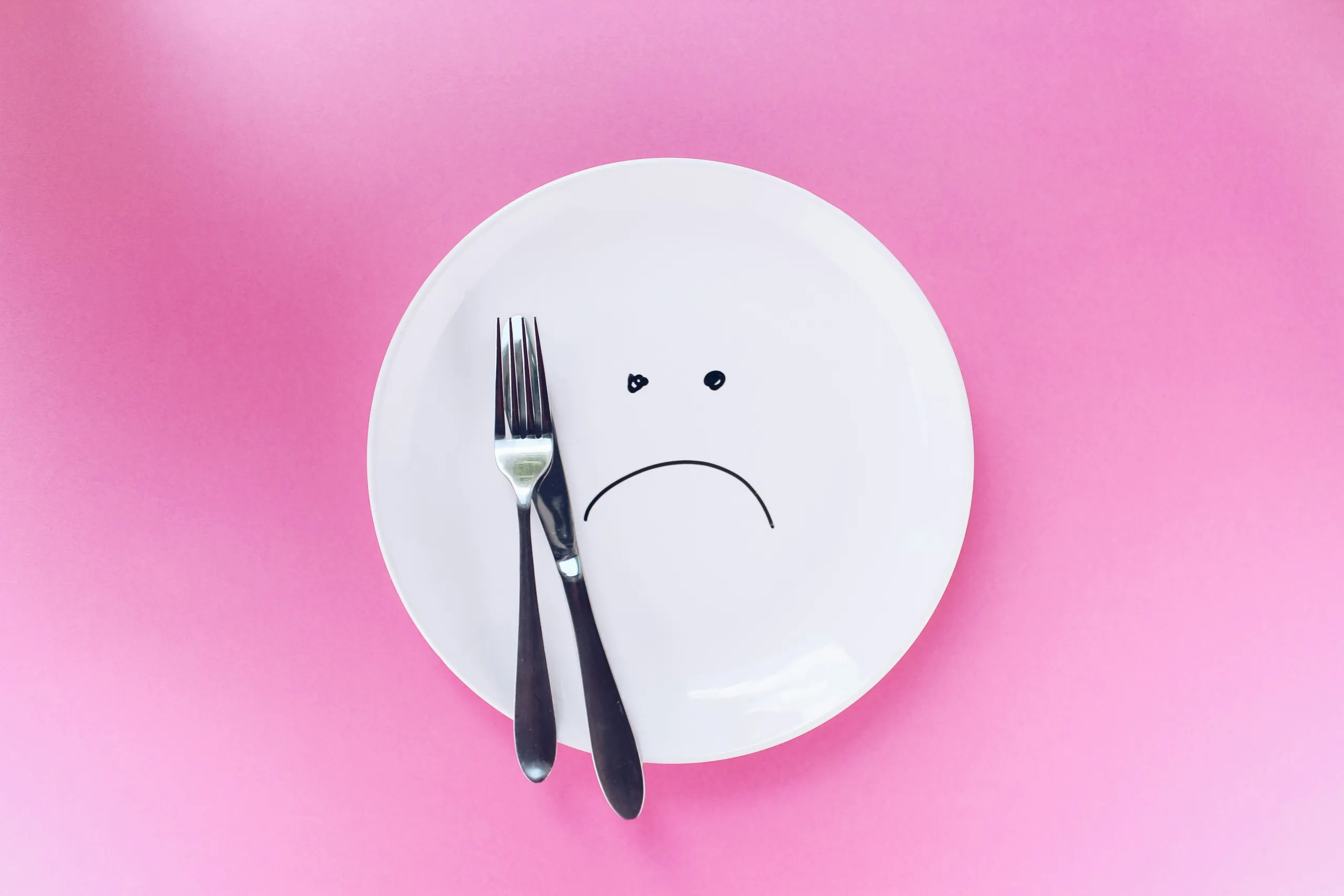 Plato blanco con una carita triste dibujada, tenedor y cuchillo sobre fondo rosa